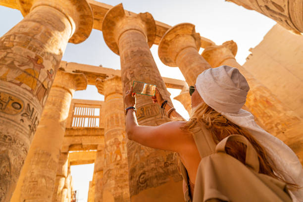 Female tourist walks between Pillars of the Great Hypostyle Hall from Karnak Temple, and taking pictures with smart phone