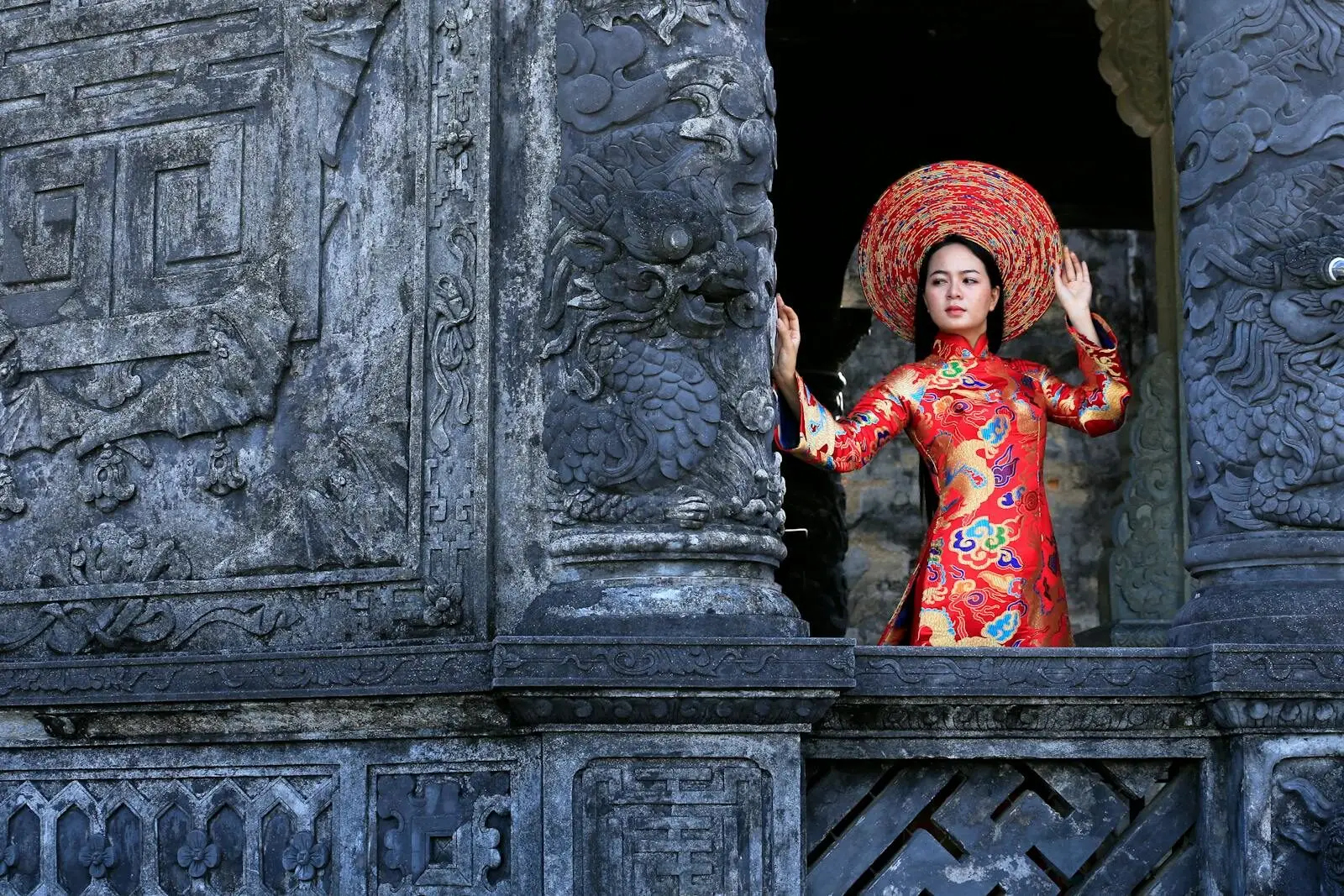 Elegant Vietnamese woman in traditional red áo dài, posing at an ancient temple with intricate stone carvings.