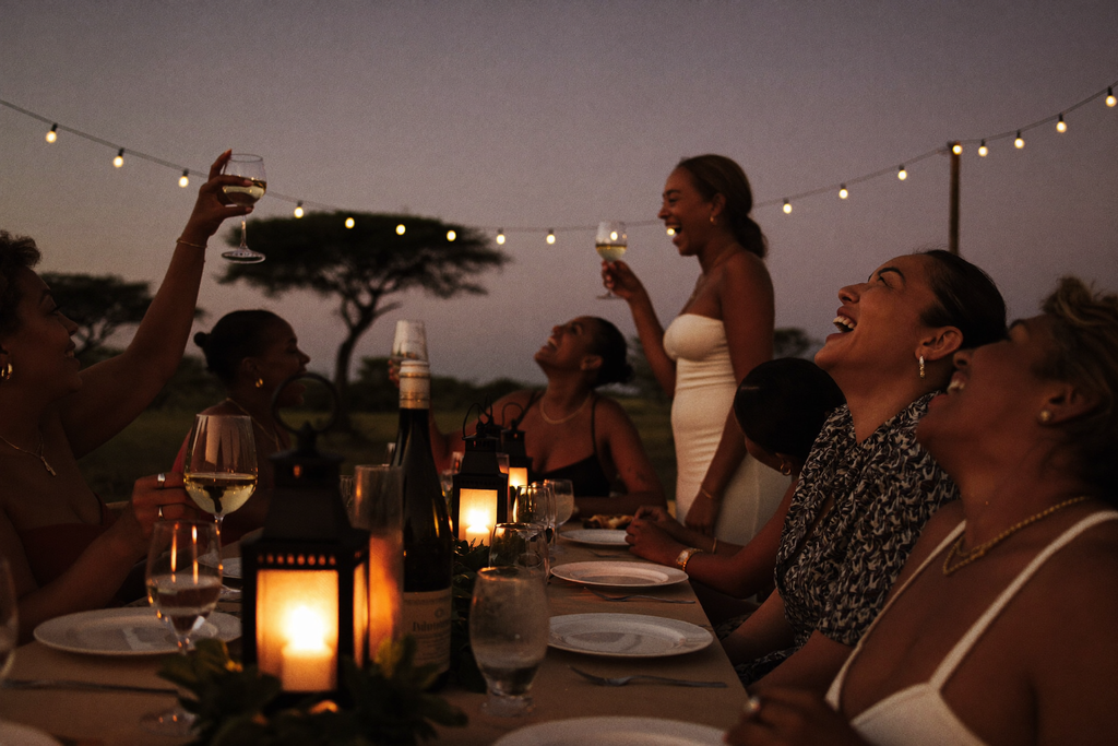 A group of seven women gathered around a lantern-lit table at an outdoor safari lodge boma at dusk, mid-toast and laughing together