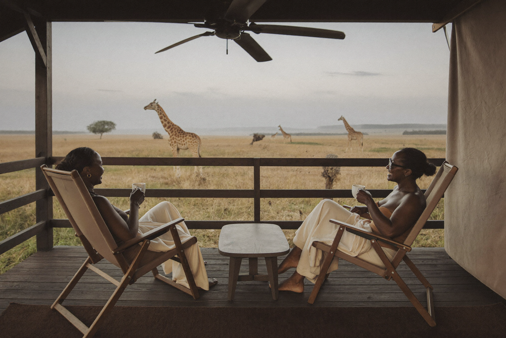 "Two women relaxed in canvas safari chairs on a private lodge veranda at sunrise, coffee in hand, looking out over a misty savanna with distant giraffes