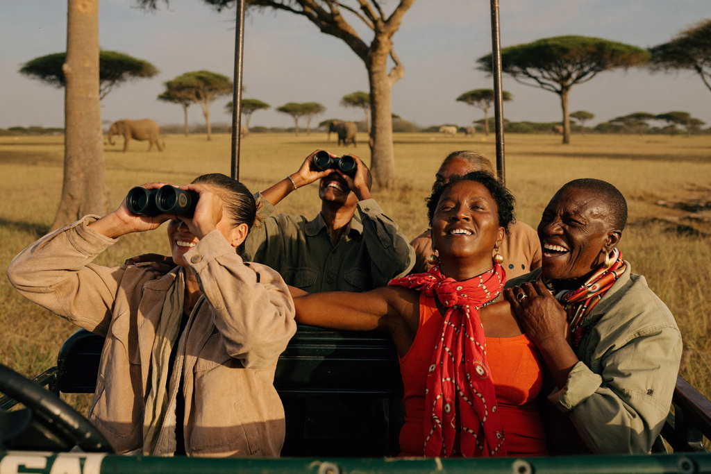 Six women in their 40s to 60s riding together in an open-sided safari vehicle at golden hour, laughing and watching elephants in the distance on the African savanna